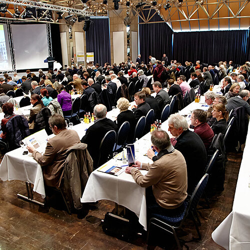 Ein Saal mit Tischen an denen Menschen sitzen mit dem Blick zur Bühne.